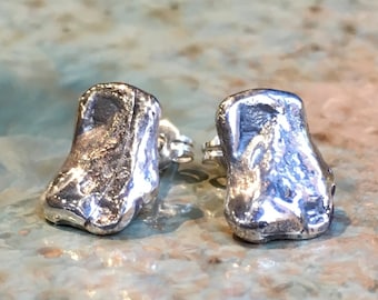 May include: A pair of silver stud earrings. Each earring has a unique, textured, organic shape. The earrings are displayed on a light blue and green surface.