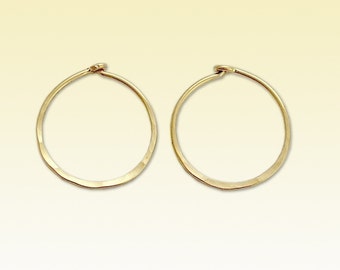 May include: A pair of gold-colored hoop earrings. The earrings are circular with a hammered texture and a simple clasp closure. The hoops are displayed against a soft, yellow background.
