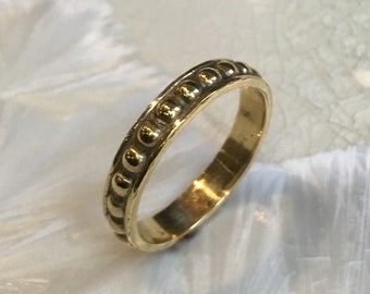 May include: A gold-colored ring with a repeating pattern of small, raised circles along the band. The ring is set against a soft, white background, highlighting its metallic sheen and intricate design. The ring appears to be a wedding band or a similar type of jewelry.