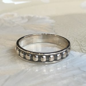 May include: A silver ring with a band of small, evenly spaced, raised circular beads. The ring has a polished, reflective surface and sits on a light-colored, textured surface. The ring is a piece of jewelry.