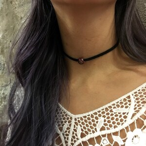 May include: A black choker necklace with a red and gold bead. The necklace is worn around the neck. The person is wearing a white lace top and has dark purple hair.
