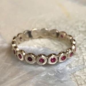 May include: A silver band ring with a row of small, round, red gemstones set in the metal. The ring is made of a smooth, polished metal.