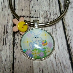 May include: A silver-colored metal binder ring with a round pendant featuring a cartoon Easter bunny holding a basket of eggs. A pink flower and yellow button accent the ring.