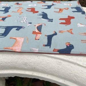 Puede incluir: Un mantel individual azul con un patrón repetido de perros de diferentes razas y colores. Los perros están ilustrados en un estilo de dibujos animados.