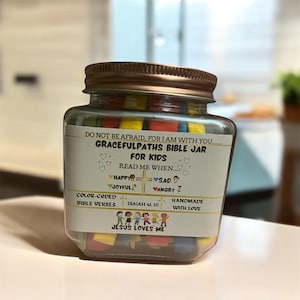 Op de afbeelding: Een glazen pot gevuld met kleurrijke vierkanten, elk met een ander Bijbelvers. De pot heeft een etiket met de tekst "Gracefulpaths Bible Jar for Kids" en bevat instructies om "Read me when..." met opties voor blij, vrolijk, verdrietig en boos. Het etiket bevat ook het vers "Isaiah 41:10" en de zin "Handmade with love".
