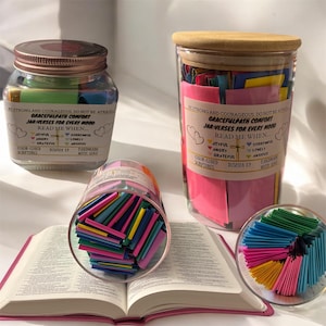 Puede incluir: Tres frascos de vidrio llenos de papeles de colores. Cada frasco tiene una etiqueta que dice "Gracefulpath Comfort Jar-Verses for Every Mood" y enumera diferentes emociones como "Joyful", "Angry", "Grateful", "Sorrowful", "Lonely" y "Anxious". Los frascos están sobre un libro encuadernado en cuero marrón.