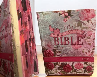 Puede incluir: Una Biblia decorativa con un diseño floral y el texto "My Creative Bible". La portada presenta rosas rosas y otras flores sobre un fondo verde claro. Las páginas están bordeadas en rosa y tienen pestañas.