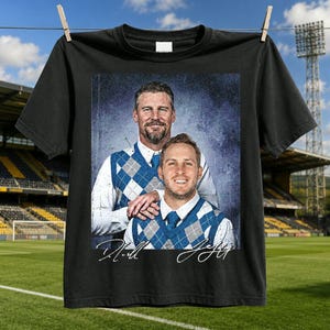 Op de afbeelding: Een zwart t-shirt met een grafische print van twee personen in witte overhemden met kraag en blauwe argyle-spencers. De print heeft een getextureerde, blauwe achtergrond in vintage-stijl en bevat twee handgeschreven handtekeningen onderaan.