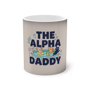 May include: A beige ceramic mug with the text "THE ALPHA DADDY" in navy blue. The mug features cartoon illustrations of a dog in a yellow floatie, a duck, and cats in a pool.