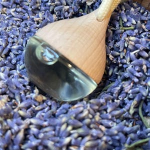 Könnte beinhalten: Ein hölzerner ätherischer Öldiffusor mit einem transparenten Glasbehälter, der an einer geflochtenen Schnur aufgehängt ist. Der Diffusor ist von getrockneten Lavendelblüten umgeben, was eine natürliche und aromatische Darstellung ergibt. Das Holz hat eine helle, natürliche Oberfläche.