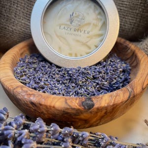 Pode incluir: Uma lata prateada com o texto "Lazy River Lavender" está em cima de uma tigela de madeira cheia de botões de lavanda secos. A tigela é rodeada por raminhos de lavanda. A lata contém um produto de cor clara.