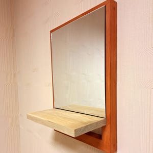 以下が含まれることがあります： 温かみのある赤茶色の木製フレームが付いた長方形の壁掛け鏡。フレームの下部には明るい色の天然木の棚が取り付けられており、外側に突き出た平らな面が小さな物を置くためのスペースとして機能しています。