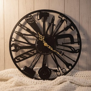 May include: Black wall clock featuring a circular design with various tools as hour markers. The clock hands are gold-colored. The tools include a saw, hammer, wrench, and pliers. The clock is set against a light wood-paneled wall.