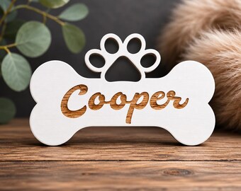 Op de afbeelding: Een witte, botvormige dierenpenning van hout met de naam "Cooper" in een bruin, gegraveerd lettertype. Een pootafdrukontwerp staat boven de naam. De tag ligt op een houten oppervlak met een vage achtergrond.