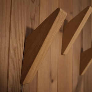 May include: Wooden wall hooks in a triangular design. The hooks are made of light-coloured wood and are mounted on a wooden wall. The hooks are angled, creating a modern and minimalist aesthetic. Suitable for coats, hats, or other items.