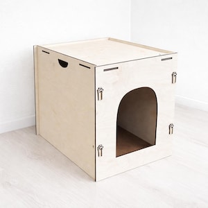 May include: A light-coloured wooden cat house with a square shape and an arched entrance. The cat house has a flat top and paw-shaped cutouts on the sides. A handle is cut into the top. The cat house is made of light-coloured wood.
