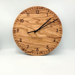 Peut inclure: Une horloge ronde en bois avec des aiguilles noires et des chiffres noirs sur un fond blanc.