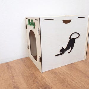 Può includere: Una cuccia per animali in legno chiaro con la sagoma ritagliata di un gatto nero che si stiracchia sul pannello laterale. Il pannello frontale presenta un ingresso ad arco e il nome Molly in corsivo verde fissato nell'angolo in alto a sinistra.