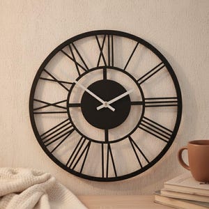 Puede incluir: Un reloj de pared redondo negro con números romanos. El reloj tiene un diseño minimalista con una esfera negra y manecillas blancas. El reloj tiene aproximadamente 46 cm de diámetro. Una taza marrón y libros están sobre una superficie de madera.