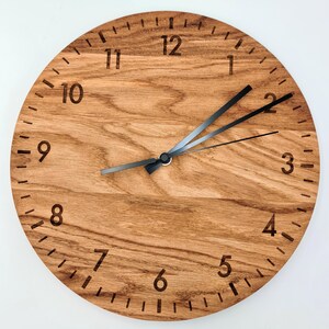 May include: A round wall clock featuring a natural light brown wood grain face with dark brown engraved numbers from 1 to 12. The clock has three thin black metal hands positioned at the center, indicating the time against a clean wood surface.