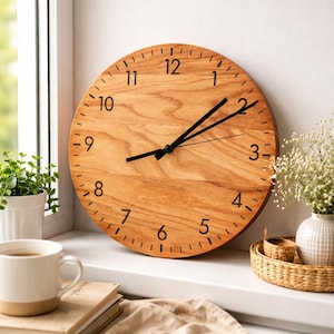 May include: A round wooden wall clock with black numerals and hands. The clock has a light brown wood grain finish and is displayed on a windowsill. A white mug, books, and a small plant are also visible.