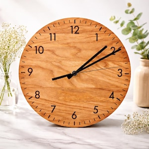 May include: A round wooden clock with a light brown finish. The clock face features black numerals and hands. The clock is sitting on a white marble surface. The clock's diameter is approximately 10 inches.