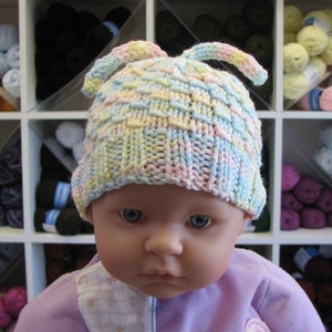 May include: A baby doll wearing a pastel pink, blue, and yellow knitted hat with ear flaps. The hat has a checkered pattern.