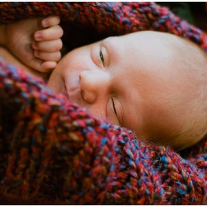 May include: A baby is sleeping soundly in a colourful knitted blanket. The baby's face is visible, with one eye closed and the other open. The blanket is a mix of red, blue and brown.