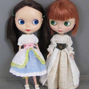 May include: Two dolls wearing dresses. The doll on the left has brown hair and blue eyes and is wearing a white and blue gingham dress with a yellow lace trim. The doll on the right has red hair and green eyes and is wearing a white dress with a brown and white gingham skirt.