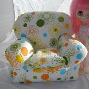 May include: A white armchair with a colorful floral pattern featuring green, yellow, blue, and orange accents. The armchair is upholstered in a soft fabric and has a rounded back and arms.