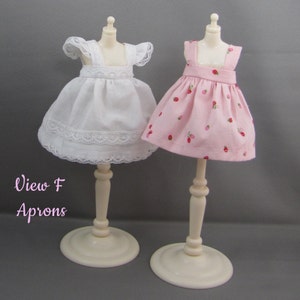 K&ouml;nnte beinhalten: Zwei Puppensch&uuml;rzen auf einer Ausstellung. Eine ist wei&szlig; mit Spitzenbesatz, die andere rosa mit Erdbeermuster. Die Sch&uuml;rzen stehen auf cremefarbenen St&auml;ndern. Der Text "View F Aprons" befindet sich unten links.