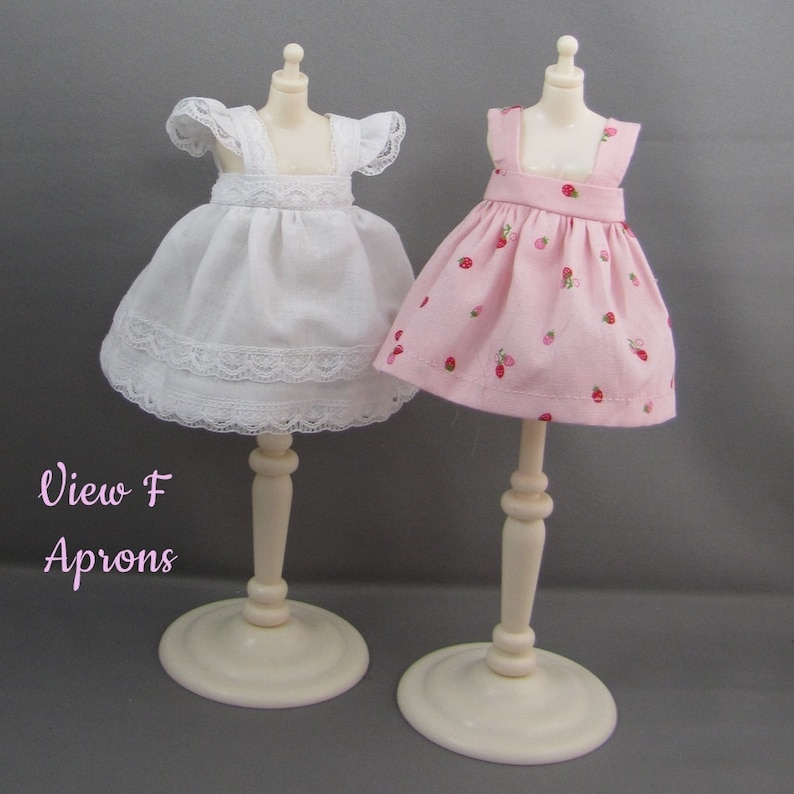 K&ouml;nnte beinhalten: Zwei Puppensch&uuml;rzen auf einer Ausstellung. Eine ist wei&szlig; mit Spitzenbesatz, die andere rosa mit Erdbeermuster. Die Sch&uuml;rzen stehen auf cremefarbenen St&auml;ndern. Der Text "View F Aprons" befindet sich unten links.