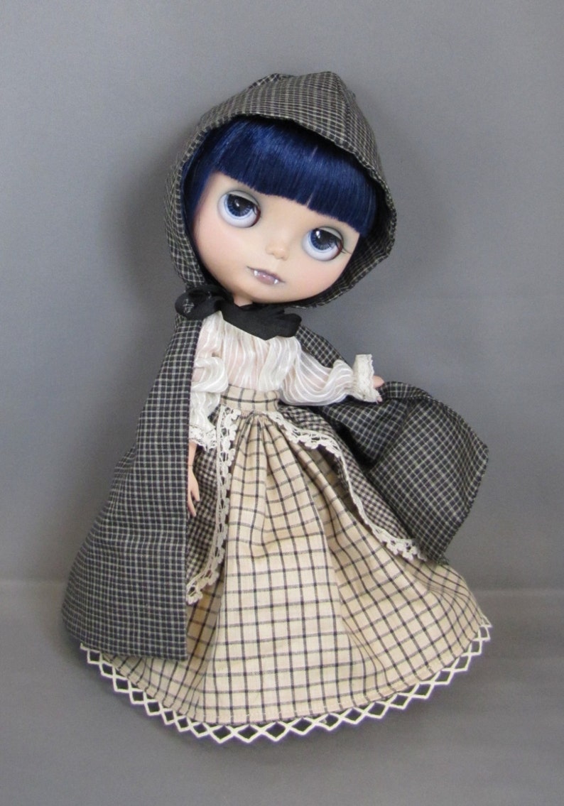 May include: A doll with blue hair and a black and white plaid dress. She is wearing a black hooded cape with a white lace trim. The doll has a painted-on expression with dark eyes and a slight smile.