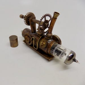 May include: A steampunk-style sculpture featuring a clear glass tube with internal components, connected to a network of copper-colored gears, pipes, and a small cylindrical piece. The intricate design showcases a blend of industrial and artistic elements.