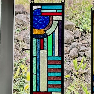 Puede incluir: Un panel rectangular de vidrieras con una composición geométrica de piezas de vidrio multicolor unidas por perfiles de plomo negros. El diseño incluye un segmento circular azul y barras horizontales y verticales en rojo, turquesa, blanco y morado.