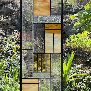Könnte beinhalten: Rechteckige Glasmalerei-Tafel mit schwarzem Rahmen. Das Design zeigt eine geometrische Anordnung verschiedener Glasstrukturen und -farben, darunter Bernstein, Klar und strukturiertes Glas. Die Tafel ist im Freien aufgehängt.