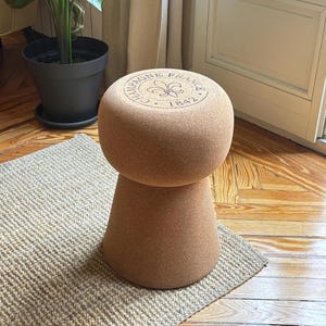May include: A cork stool shaped like a champagne bottle stopper. The top of the stool has the text "Champagne France 1842" and a fleur-de-lis design.