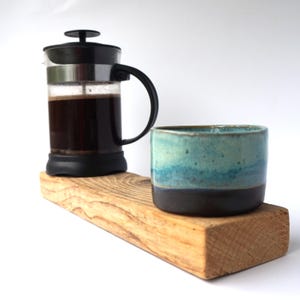 May include: A French press coffee maker with a black handle and lid, filled with dark coffee, sits on a wooden plank. A handmade ceramic mug with blue and teal glaze and a dark brown base is next to it. The items are on a white background.