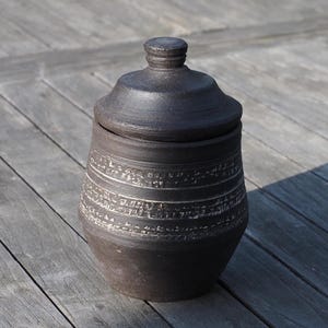 May include: A dark brown ceramic jar with a lid. The jar has a rounded body and a lid with a small knob. The jar is decorated with horizontal bands of etched designs. The jar is sitting on a wooden surface.