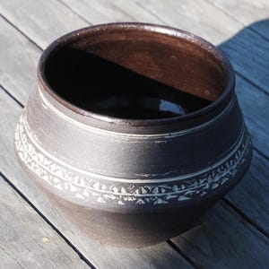 Könnte beinhalten: Eine dunkelbraune Keramikschale mit einem dekorativen weißen Muster in der Mitte. Die Schale hat eine weite Öffnung und steht auf einer verwitterten Holzoberfläche. Das Innere der Schale ist ein dunklerer Braunton.