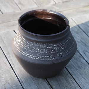 Könnte beinhalten: Ein dunkelbrauner Keramiktopf mit einem breiten, abgerundeten Körper und einem leicht ausgestellten Rand. Der Topf weist in der Mitte ein Band aus komplizierten, geometrischen Mustern in einer helleren Farbe auf. Der Topf steht auf einer Holzoberfläche.