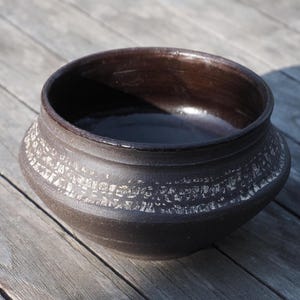 Könnte beinhalten: Eine dunkelbraune Keramikschale mit breiter, runder Form. Die Schale hat ein Band aus weißen, strukturierten Details in der Mitte. Das Innere ist glänzend dunkelbraun und steht auf einer Holzoberfläche.