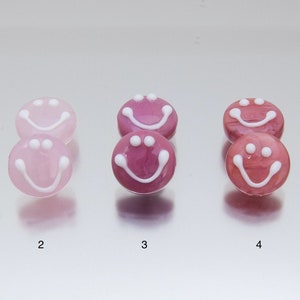 May include: Five pairs of glass smiley face earrings in various shades of pink and purple. The earrings are arranged in a row from lightest to darkest.