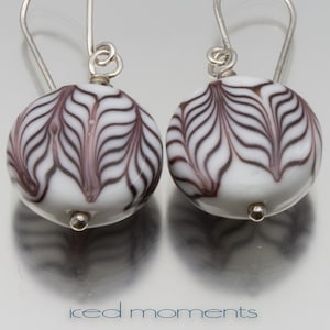 May include: A pair of round white earrings with a brown and purple swirl design. The earrings have silver-colored hooks and small silver-colored beads at the bottom. The text "iced moments" is at the bottom.