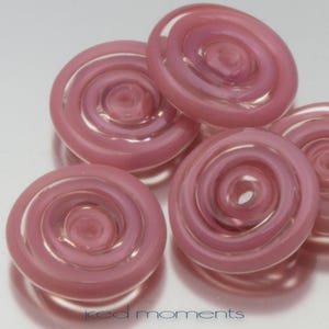 May include: Five pink glass buttons with a swirl design. The buttons are translucent and have a slight iridescent sheen.