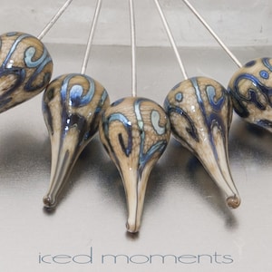 May include: Five teardrop-shaped glass beads with a brown and blue swirl pattern are attached to silver pins. The beads are arranged in a semi-circle.