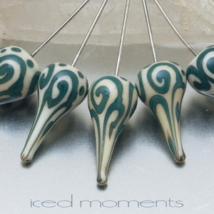 May include: Five white teardrop-shaped beads with a green swirl pattern are attached to silver pins. The beads are arranged in a fan shape. The text "iced moments" is printed below the beads.