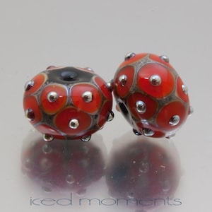 May include: Two red and black glass beads with silver accents. The beads have a geometric pattern of circles and are designed to look like ladybugs.