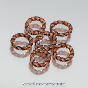 Könnte beinhalten: Sechs dekorative Glasringe mit einem gedrehten Muster in Braun-, Orange- und Schwarztönen. Die Ringe sind auf einer reflektierenden Oberfläche angeordnet, mit dem Text "iced moments" unten.