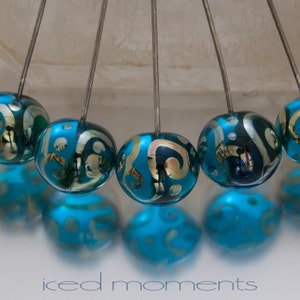 Op de afbeelding: Een close-up van een blauwe en gouden glazen kraal die aan een draad hangt. De kraal is opgehangen aan een draad en maakt deel uit van een mobiel. De mobiel hangt aan een plafond.